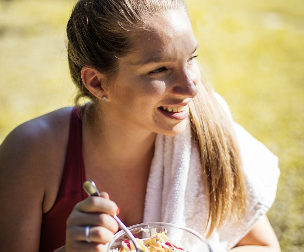smiling girl with a blonde ponytail and a towel over her shoulder outside in the park eating a health snack